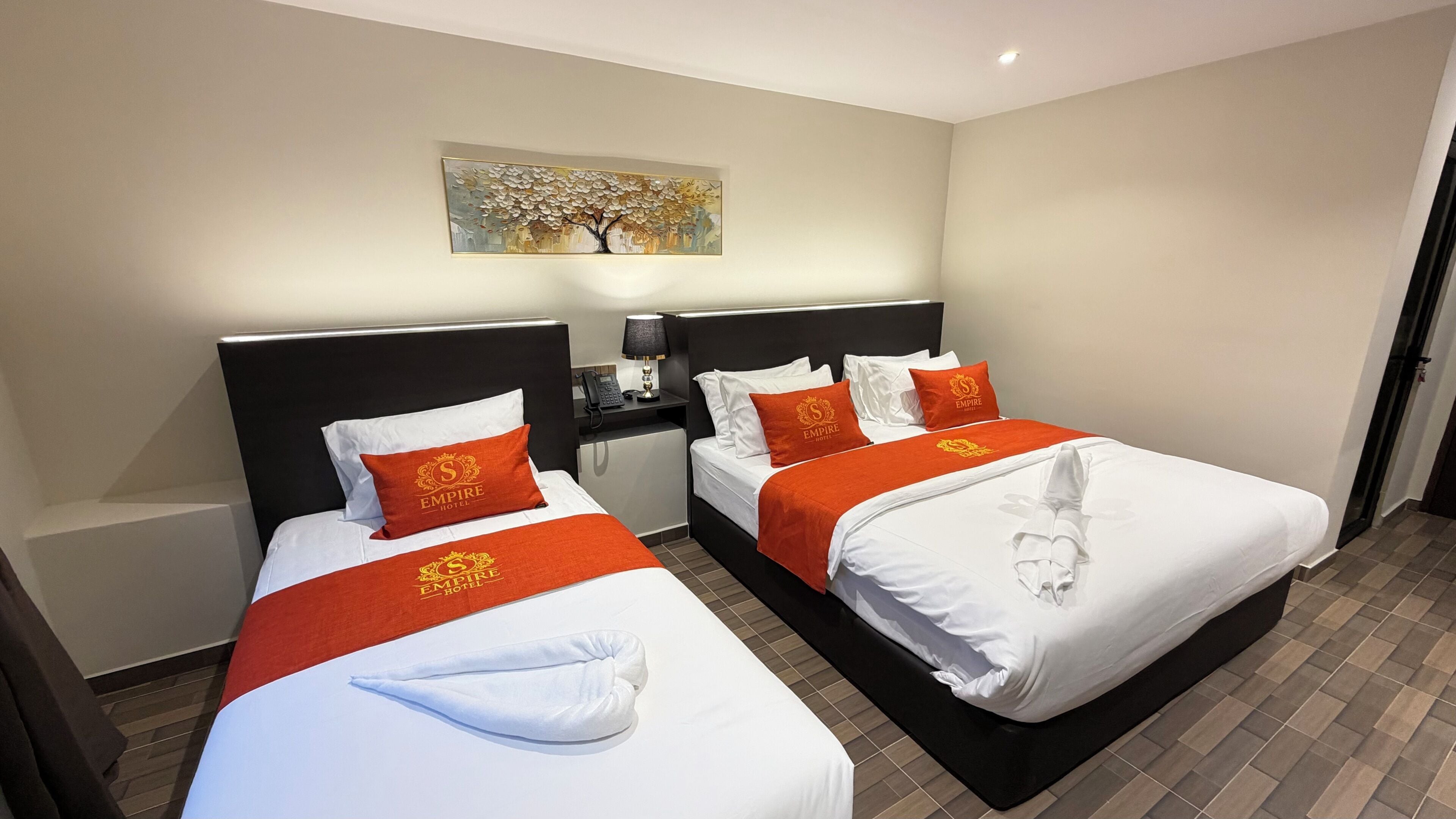 Habitación de hotel con dos camas, cada una con ropa de cama blanca y detalles anaranjados, un cuadro en la pared y una mesita de noche con lámpara.