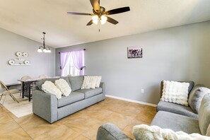 A living room with a gray sofa, a dining table with chairs, a ceiling fan, and a wall-mounted light fixture. - Pool, Hot Tub & Kids' Play Area! Davenport Home (Davenport)