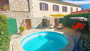 Una piscina de piedra con escalera, rodeada por un patio de piedra, una silla de descanso y una mesita.
