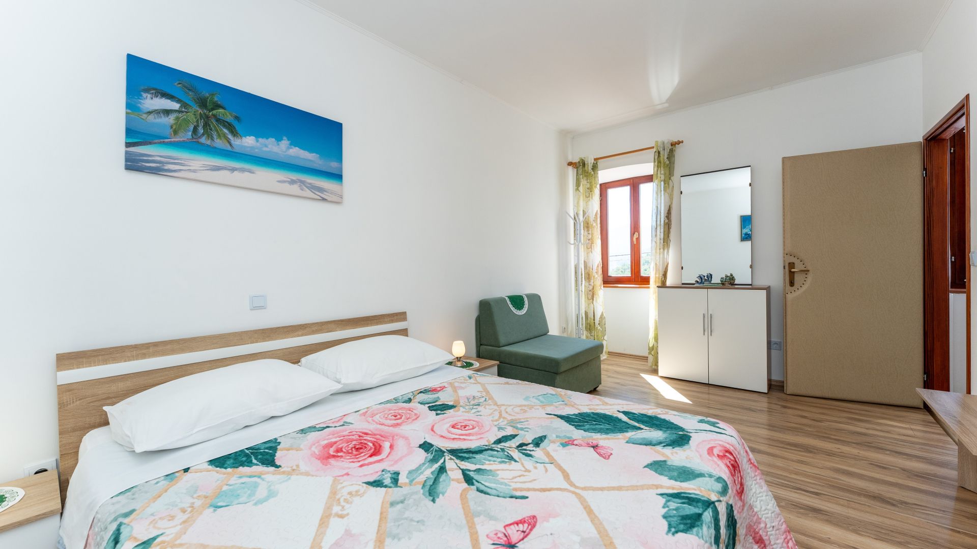 A hotel room with a bed, a chair, a wardrobe, and a painting of a beach scene.