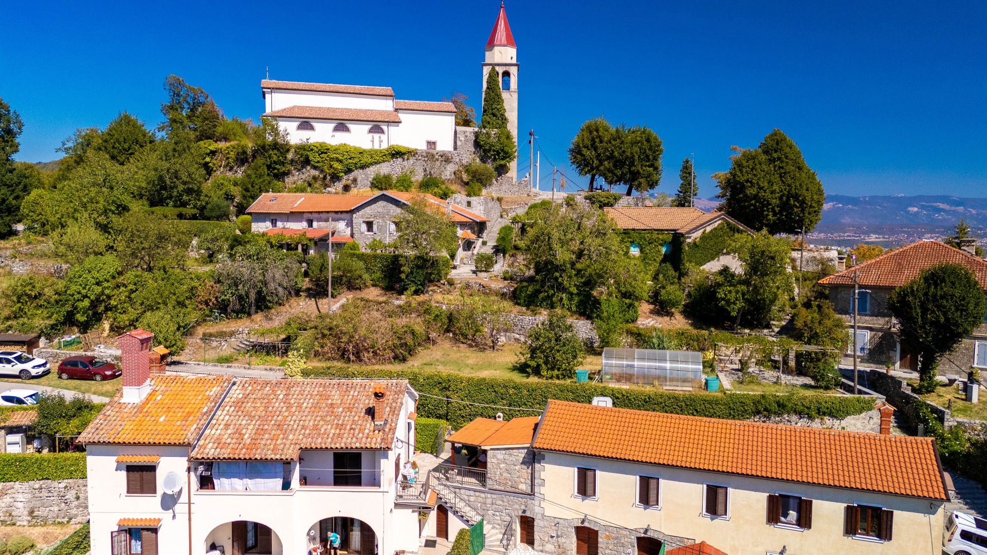 Ein Dorf an einem Hang mit traditionellen Steinhäusern, einer Kirche mit Glockenturm und weiter Aussicht auf die umliegende Landschaft.
