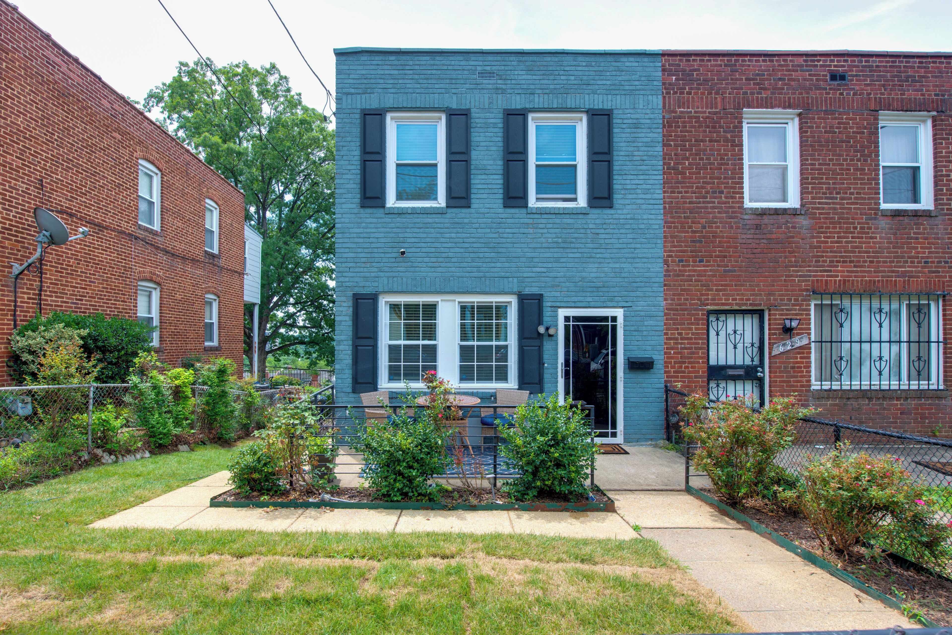 A two-story residential building with a blue exterior, a front door, and a small front yard with plants and a patio.
