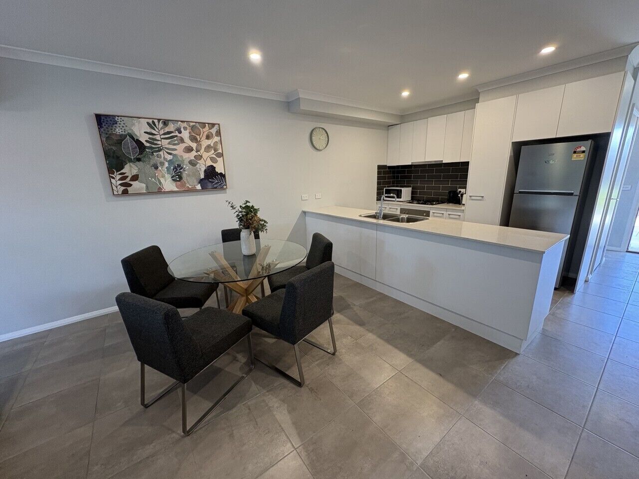 A modern kitchen with white cabinets, a stainless steel refrigerator, and a dining area with a glass table and four chairs.