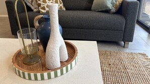 Uma sala de estar com um sofá cinzento, almofadas estampadas, um vaso e um copo sobre um tabuleiro de madeira.