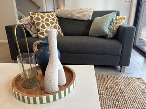 Un salon avec un canapé gris, des coussins à motifs, un vase et un verre posés sur un plateau en bois.