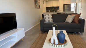 Uma sala de estar moderna com uma televisão de tela plana em um suporte branco, um sofá cinza com almofadas estampadas e uma mesa de centro com vasos decorativos.
