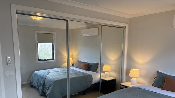 Quarto com roupeiro de portas deslizantes, duas camas, abajures e uma janela.