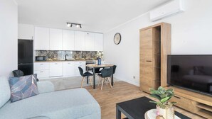 Un menjador modern amb un sofà, una zona de dinar amb taula i cadires, una cuina amb armaris blancs, i un televisor sobre un moble de fusta.