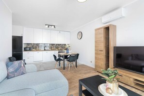 Un menjador modern amb un sofà, una zona de dinar amb taula i cadires, una cuina amb armaris blancs, i un televisor sobre un moble de fusta.