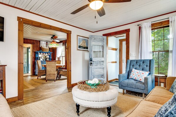 A cozy living room with a wooden ceiling, a dining area with wicker furniture, and a large window with curtains. - Tiny Antique Cottage - 2 Minutes From Short Sands! (York)