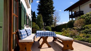 Ein Sitzbereich im Freien mit zwei hölzernen Bänken und einem Tisch, einer Fußmatte sowie einem Gebäude mit grüner Tür und Fenstern.