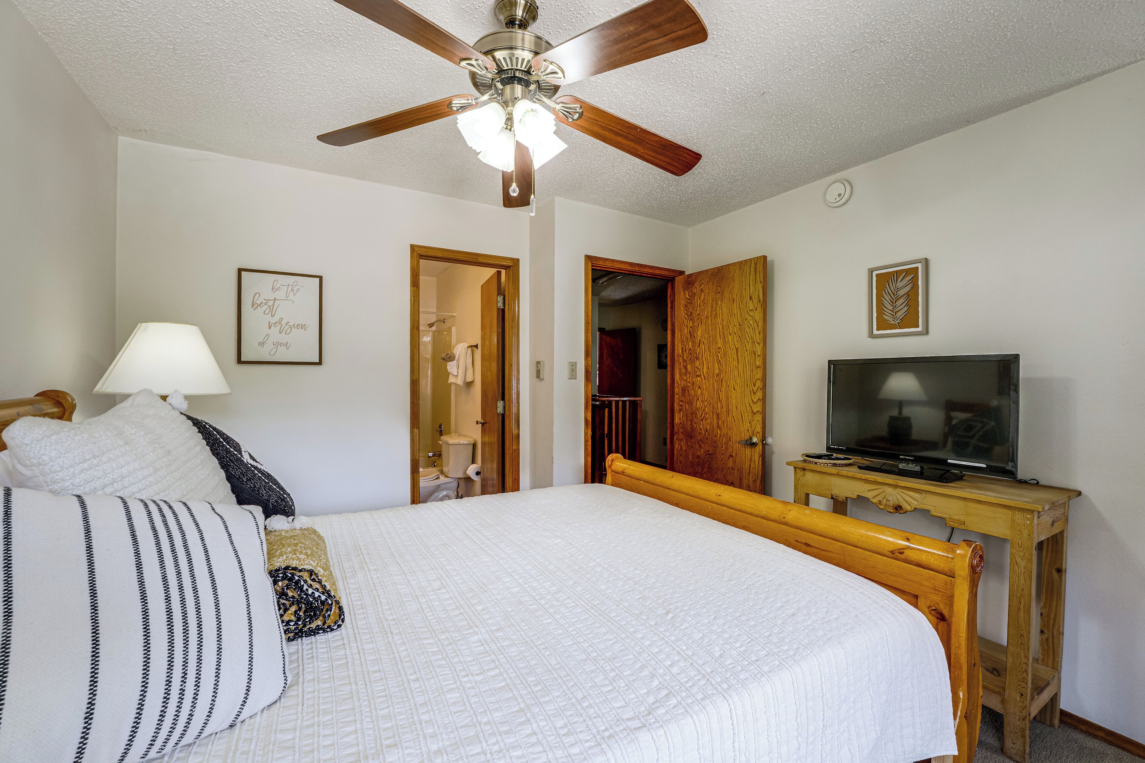 A bedroom with a bed, a television on a wooden stand, a ceiling fan, and a door leading to a bathroom.