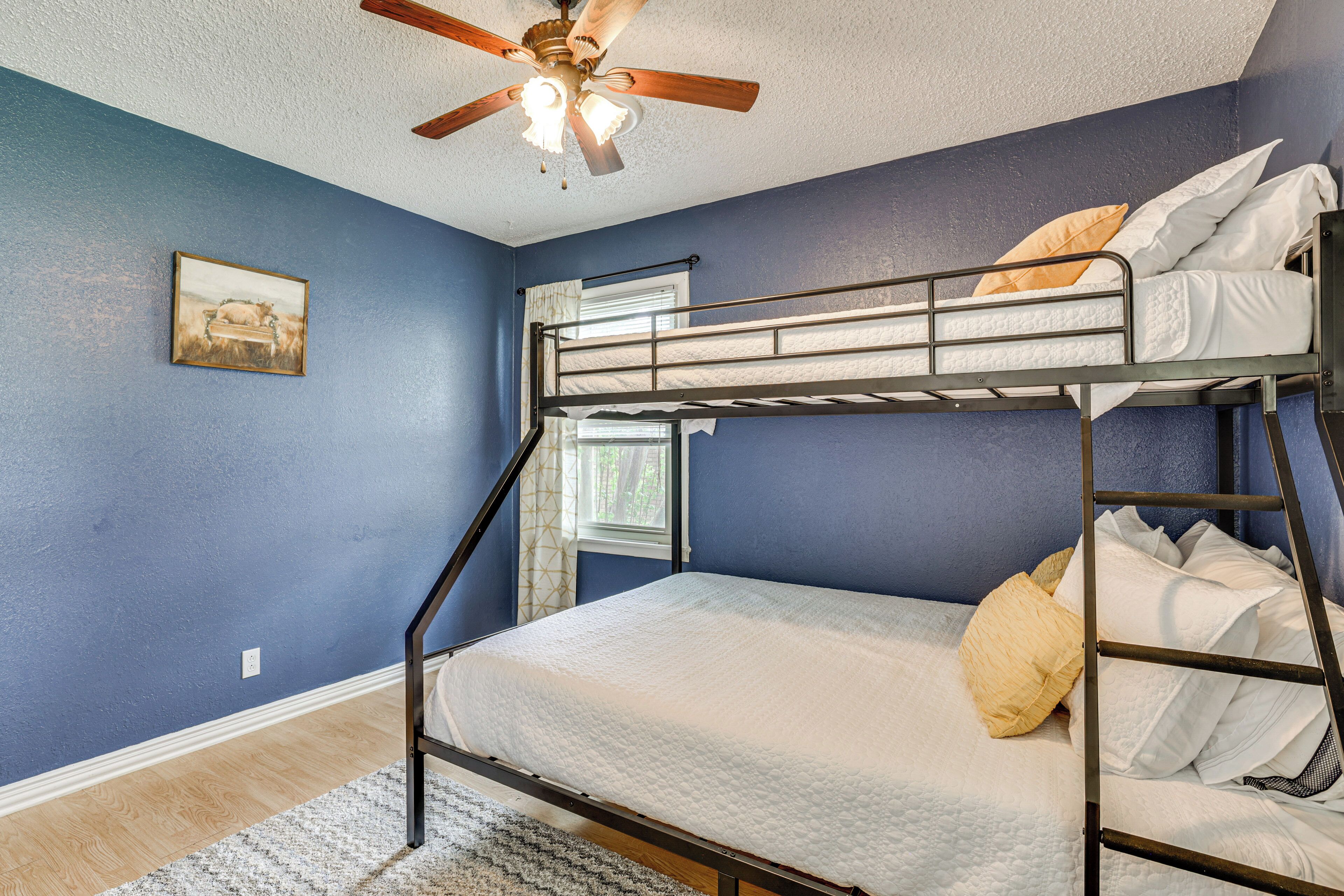 Une chambre avec un lit superposé, un ventilateur de plafond, une fenêtre avec des rideaux et un tableau accroché au mur.