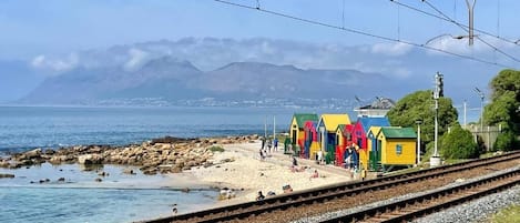 Vías de tren costeras con cabañas de playa coloridas y montañas al fondo.