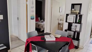 Un coin repas avec une table, des chaises et une nappe rouge. On y trouve une bibliothèque remplie de divers objets et une porte qui donne sur une autre pièce.
