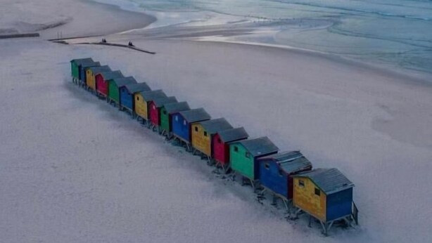 A row of colorful beach huts on a sandy beach with a view of the ocean and distant mountains.