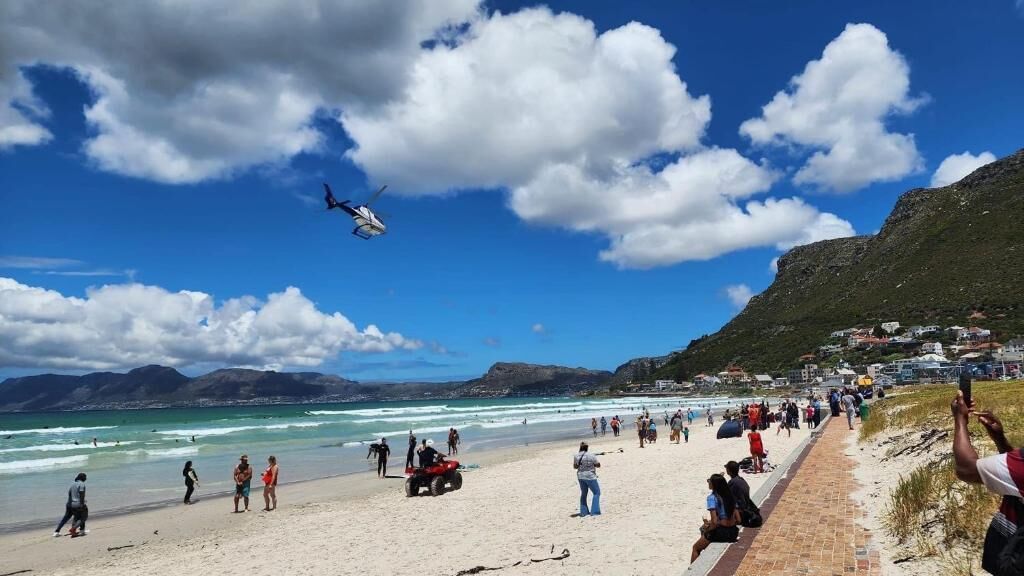 A beach with a helicopter, people, and a coastal town in the background.