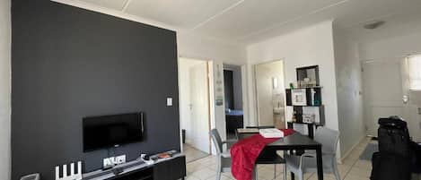 Un salon moderne avec un téléviseur à écran plat, une table à manger avec des chaises et, au fond, un coin cuisine.