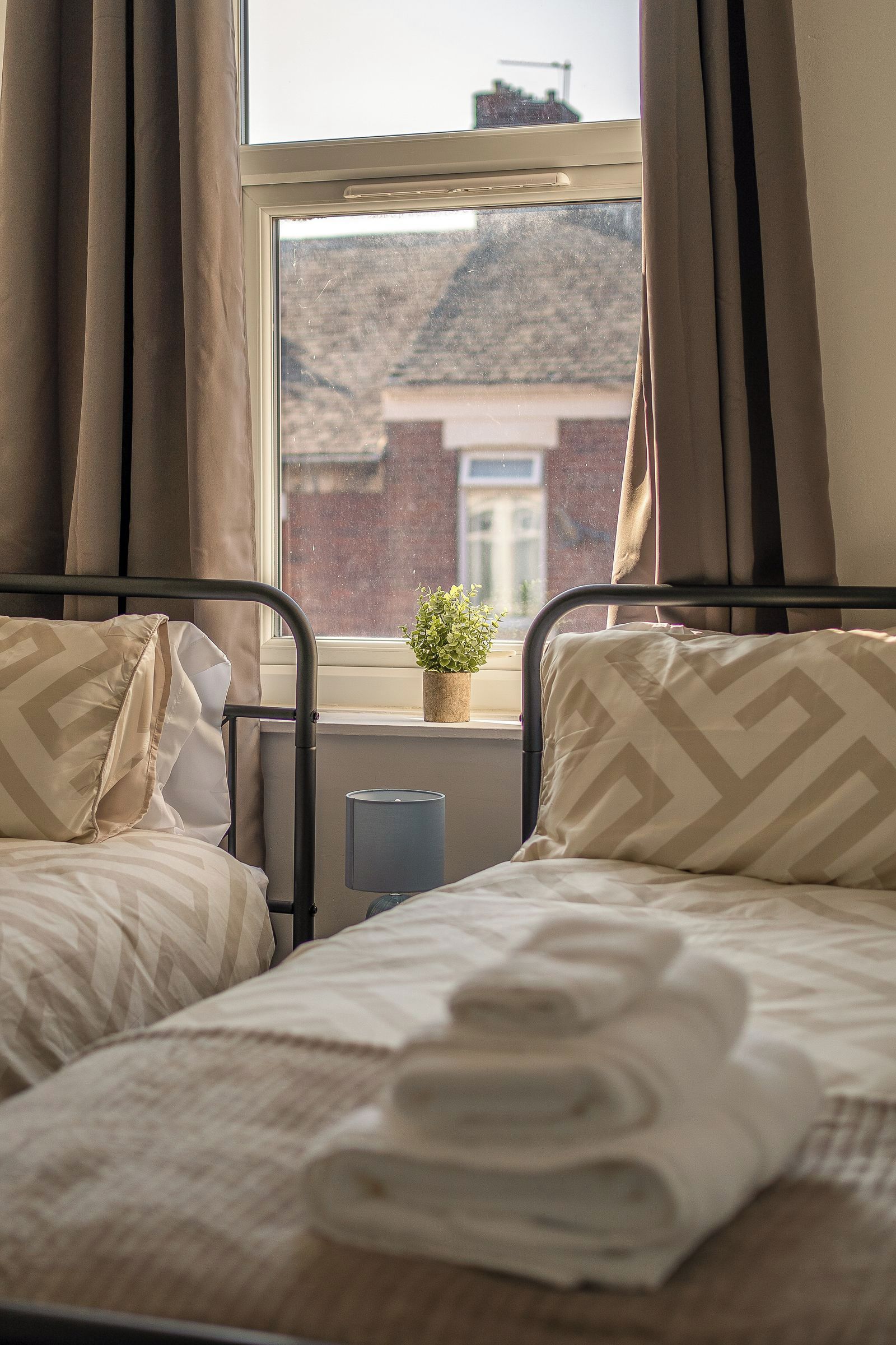 A bedroom with two beds, a window with curtains, a radiator, and a small plant on the windowsill.