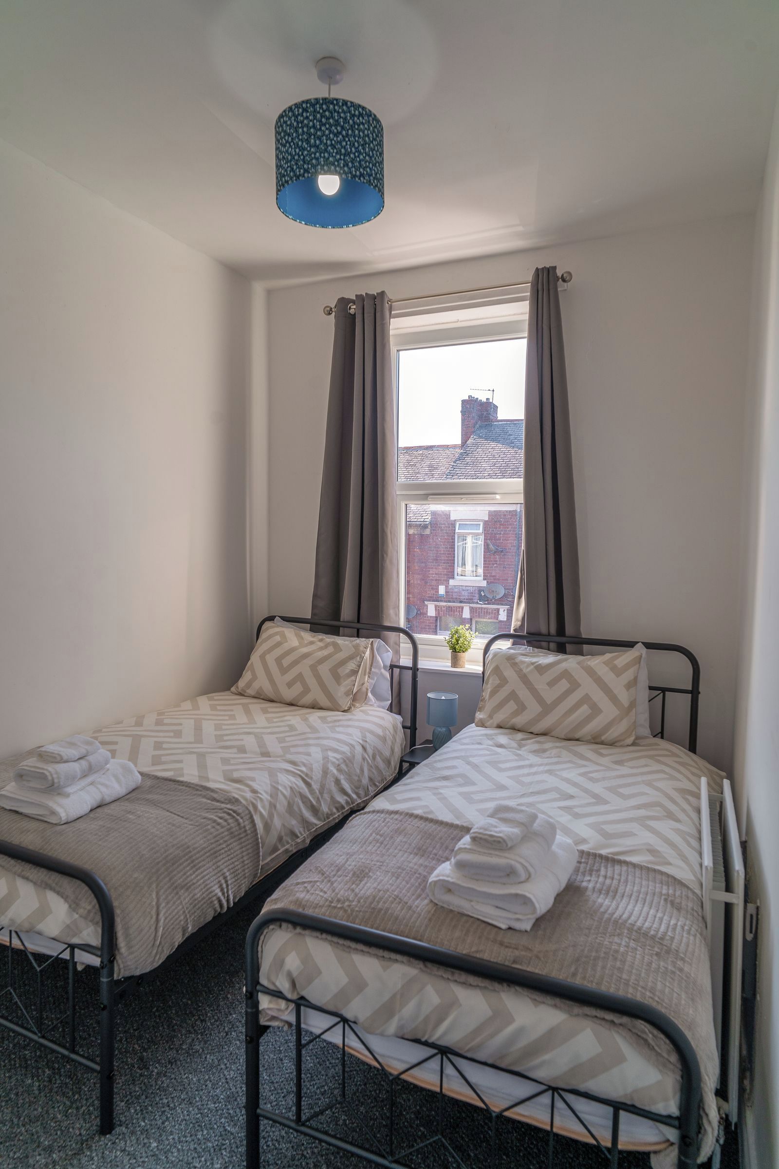 Une chambre avec deux lits simples, une fenêtre avec des rideaux, une petite table avec une lampe et une vue sur un bâtiment à l’extérieur.