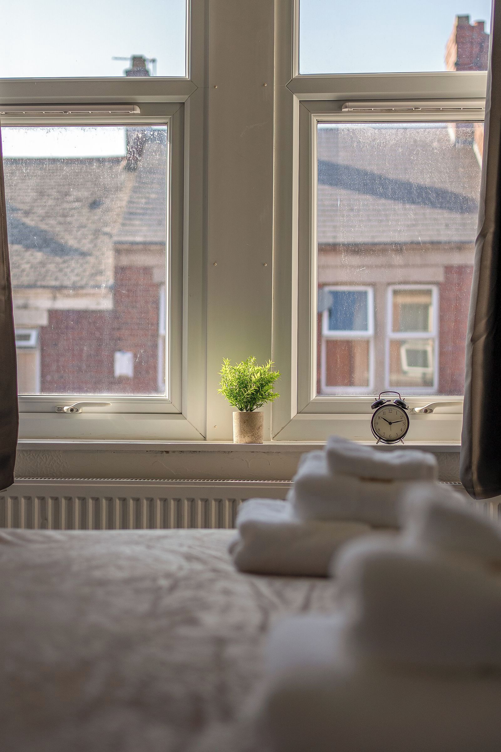 Ein Schlafzimmer mit zwei Fenstern, einer kleinen Topfpflanze und einem Nachttisch mit einer Uhr.