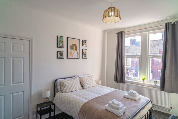 Een slaapkamer met een bed, een nachtkastje met een lamp, een raam met gordijnen en decoratieve muurkunst.