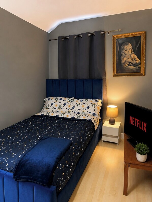 A bedroom with a bed, a nightstand with a lamp, a TV displaying 'NETFLIX,' and a framed picture on the wall.