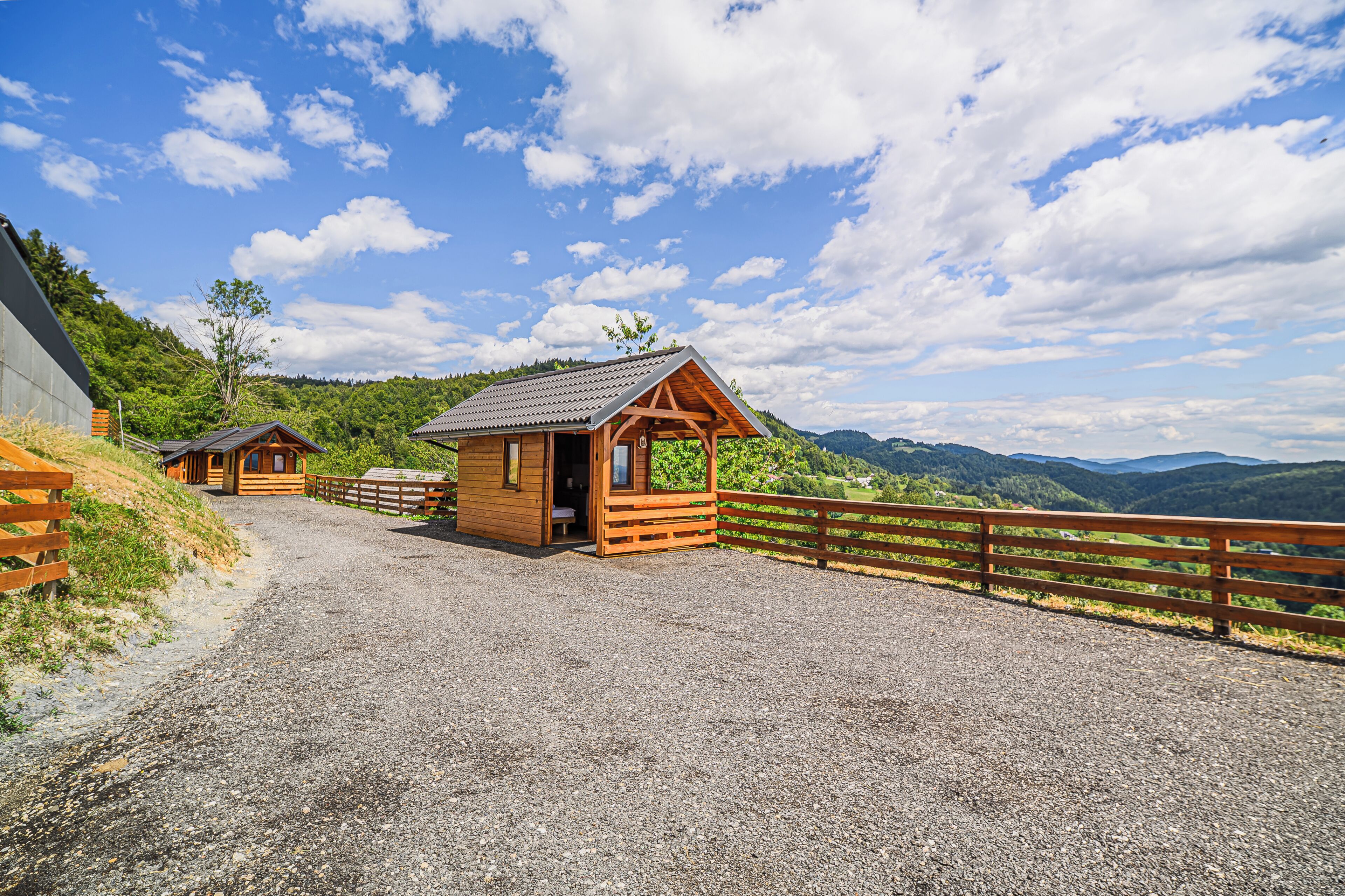 A wooden cabin with a gravel driveway and a wooden fence, surrounded by a mountainous landscape.