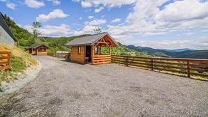 Una cabaña de madera con camino de entrada de grava y cerca de madera, rodeada por un paisaje montañoso.