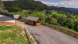 Un paisaje rural con una pequeña construcción de madera, una zona cercada y vistas a un valle con casas y árboles.