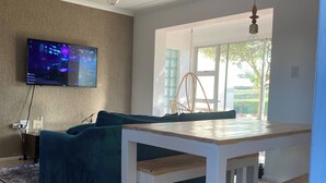 Un salon moderne avec un téléviseur à écran plat, un canapé bleu, une table à manger en bois et un luminaire suspendu.