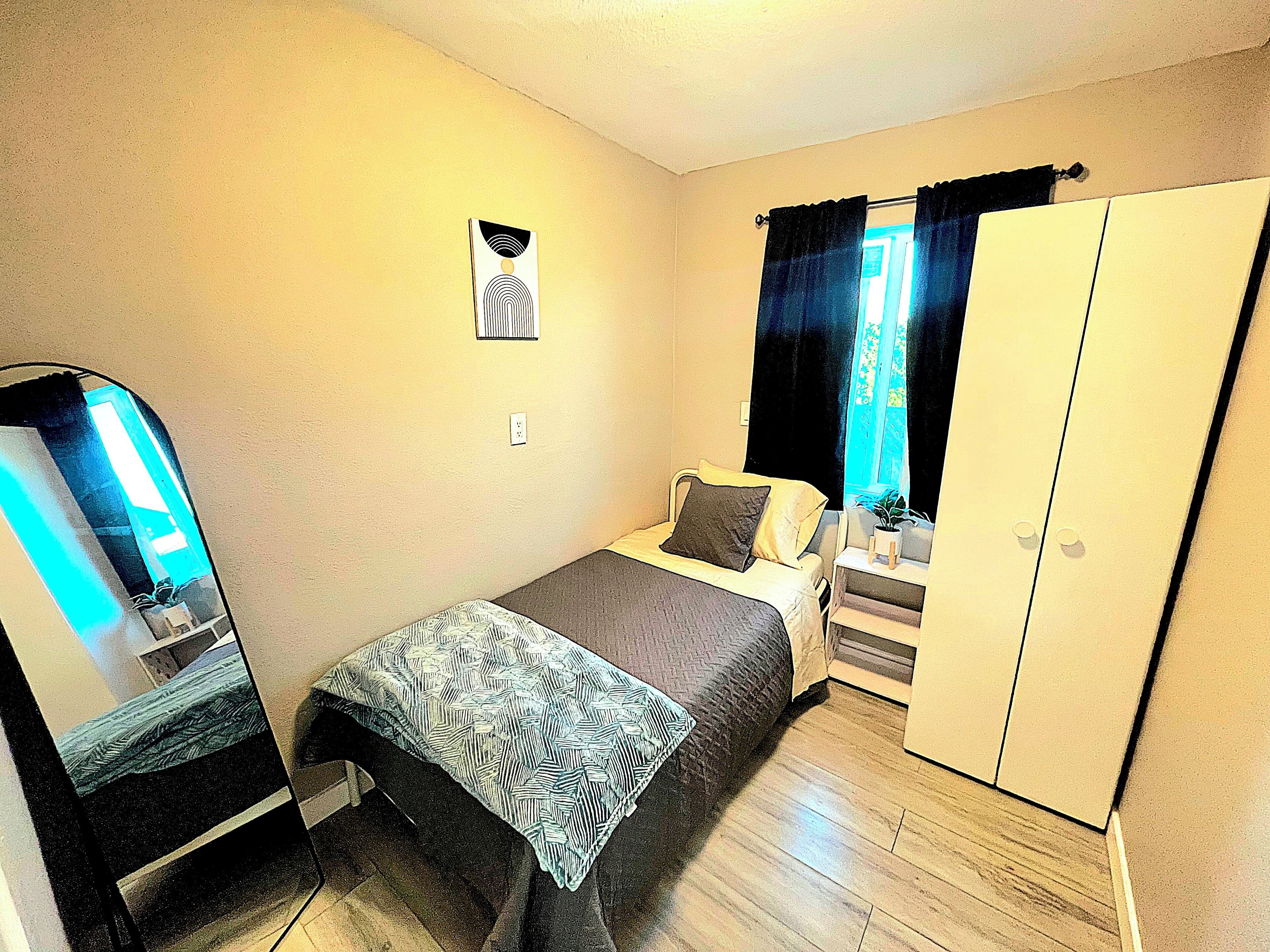 Ein kleines Zimmer mit einem Einzelbett, einem Kleiderschrank, einem Spiegel und einem fenster mit Vorhängen.