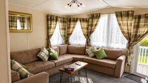 A living room with a beige sectional sofa, a wooden coffee table, and plaid curtains. - Aldous Rest - Cosy Park Home in quiet Norfolk Countryside (Fritton)