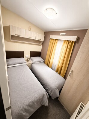 A compact room with two beds, a small bedside table, a ceiling light, and a window with curtains.