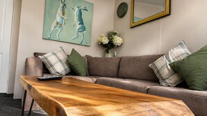 A living room with a wooden coffee table, a sofa with plaid cushions, a painting of rabbits, a vase with flowers, and a mirror on the wall.