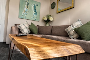 Un salón con una mesa de centro de madera, un sofá con cojines escoceses, un cuadro de conejos, un jarrón con flores y un espejo en la pared.