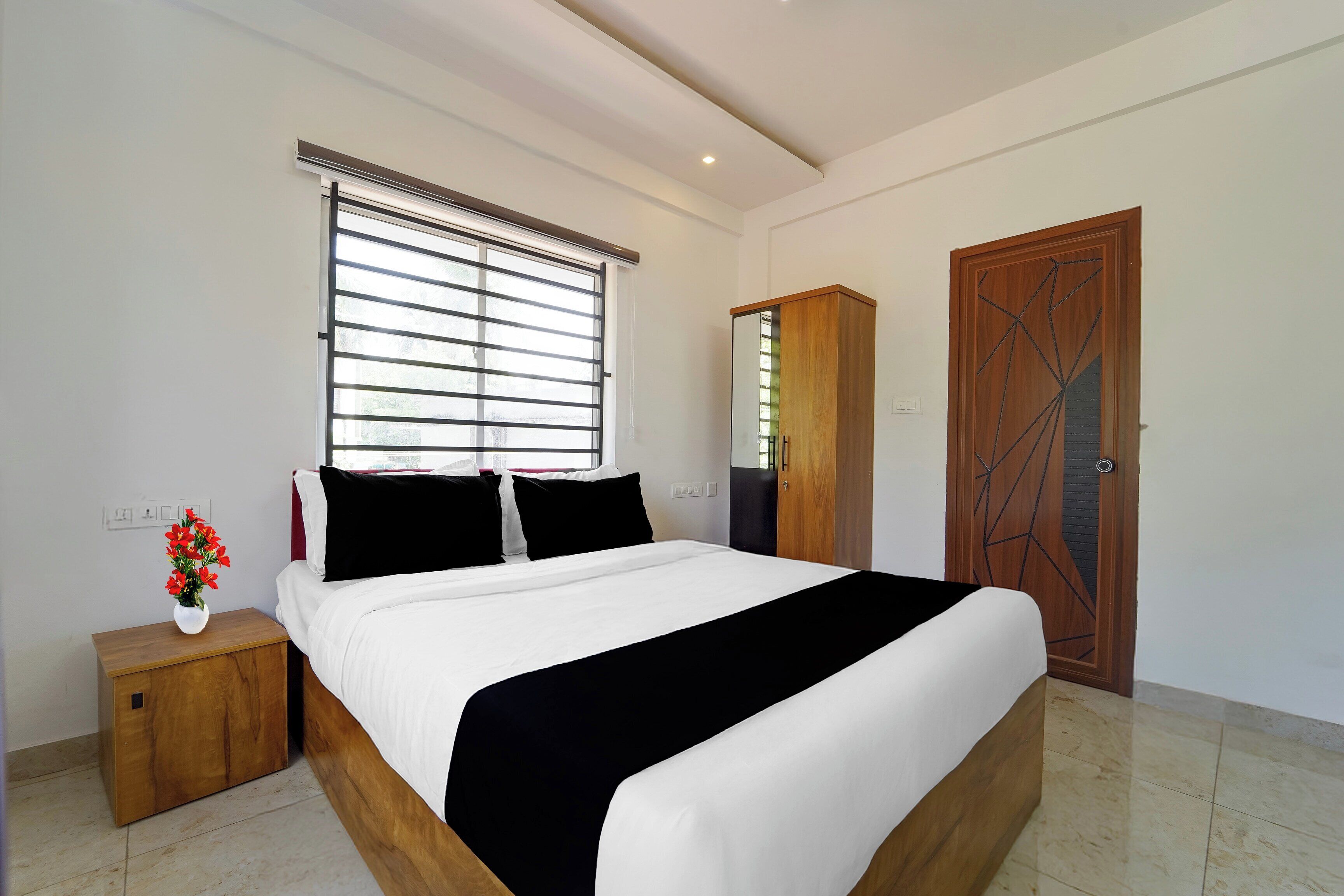 Un dormitorio con una cama, una mesita de noche, un armario de madera, una puerta con diseños geométricos y una ventana con persianas.