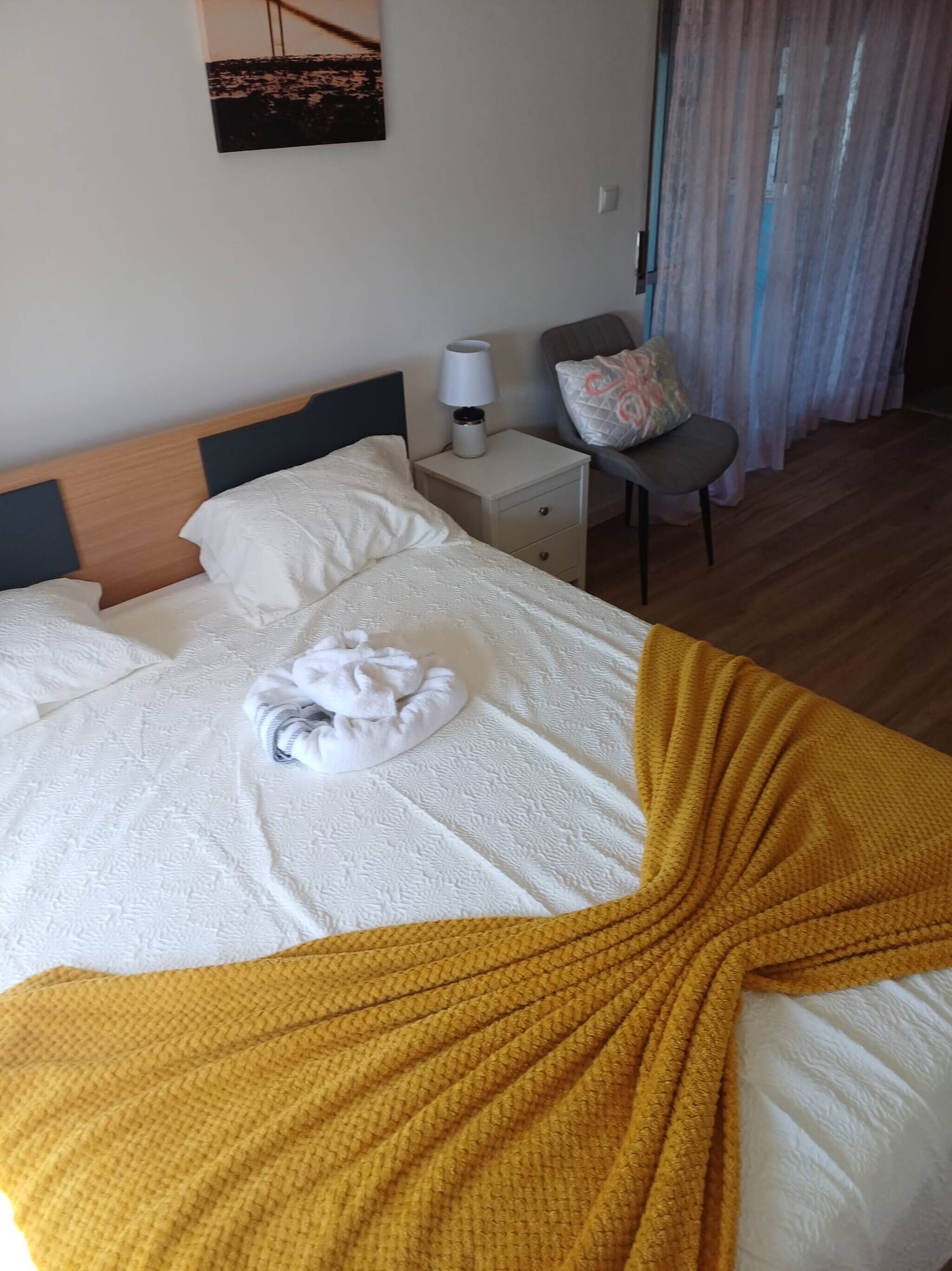 A neatly made bed with a white duvet, a yellow throw blanket, and a towel on the bed. There is a nightstand with a lamp and a pillow with a floral pattern. A chair with a cushion is visible in the background.