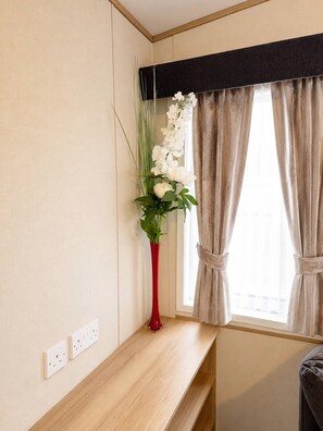 A room with a wooden bench, a red vase with white flowers, and a window with beige curtains.
