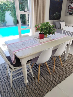 Ein Essbereich mit Tisch für vier Personen, weißen Stühlen, einem Blumentopf und Blick auf einen Swimmingpool.