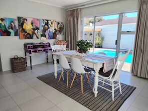 A dining area with a table and chairs, a bookshelf, and a view of a swimming pool through large windows. - Cornerhouse Family Retreat (Cape Town)