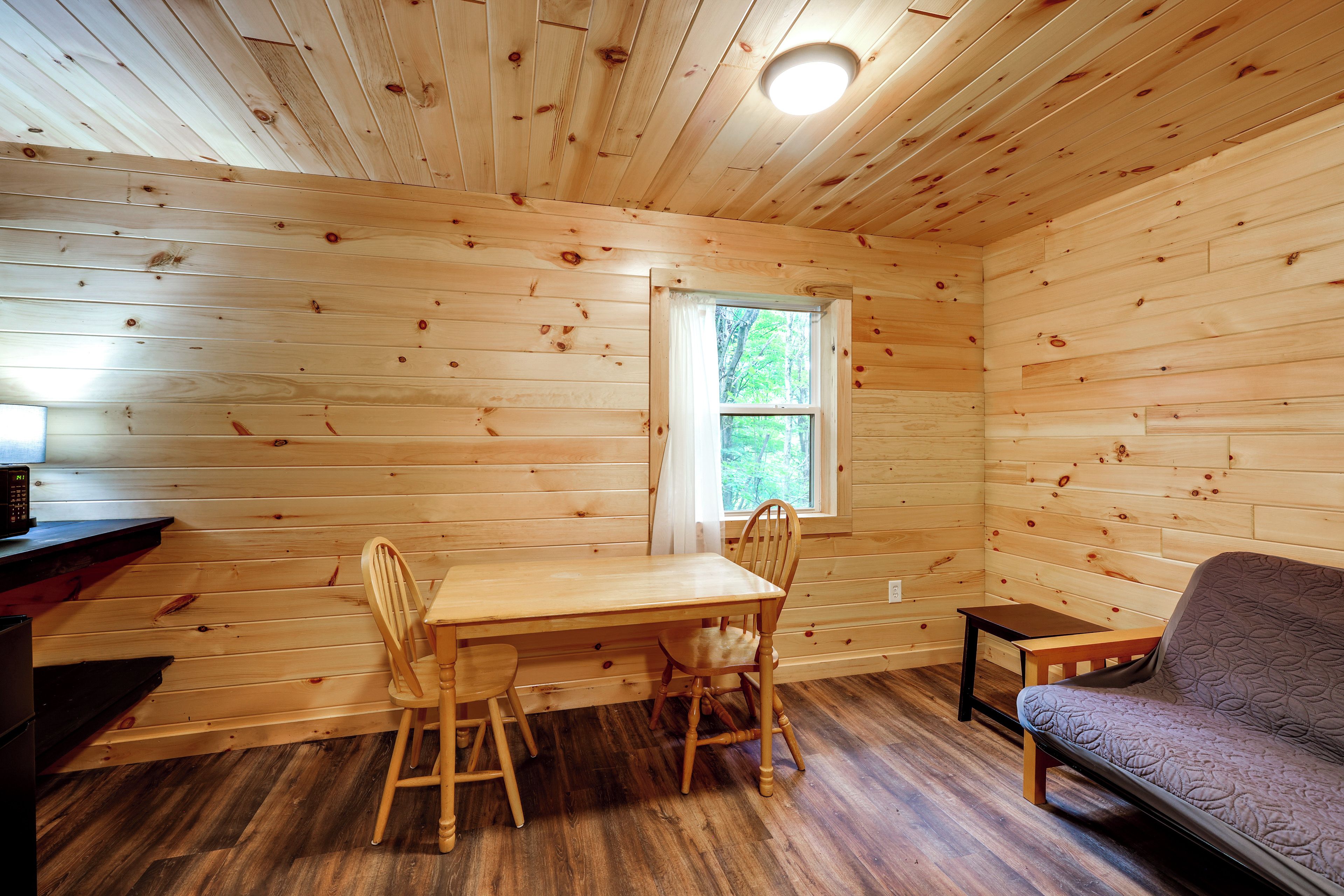 A wooden cabin interior with a dining table, chairs, a small table, a sofa, and a window with a view of trees. - Letchworth Cabin: Lake Access, Slide, Canoe Rental (Bliss)