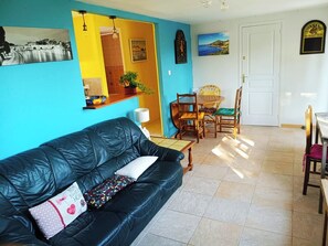 Ein Wohnzimmer mit schwarzem Ledersofa, einem Esstisch mit Stühlen, einer Küchenzeile mit Spüle und Arbeitsfläche sowie einer Tür zu einem weiteren Raum.