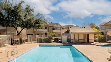 Zona de piscina con cielo despejado, piscina cercada, sillas de descanso y un edificio con zona de estar cubierta.