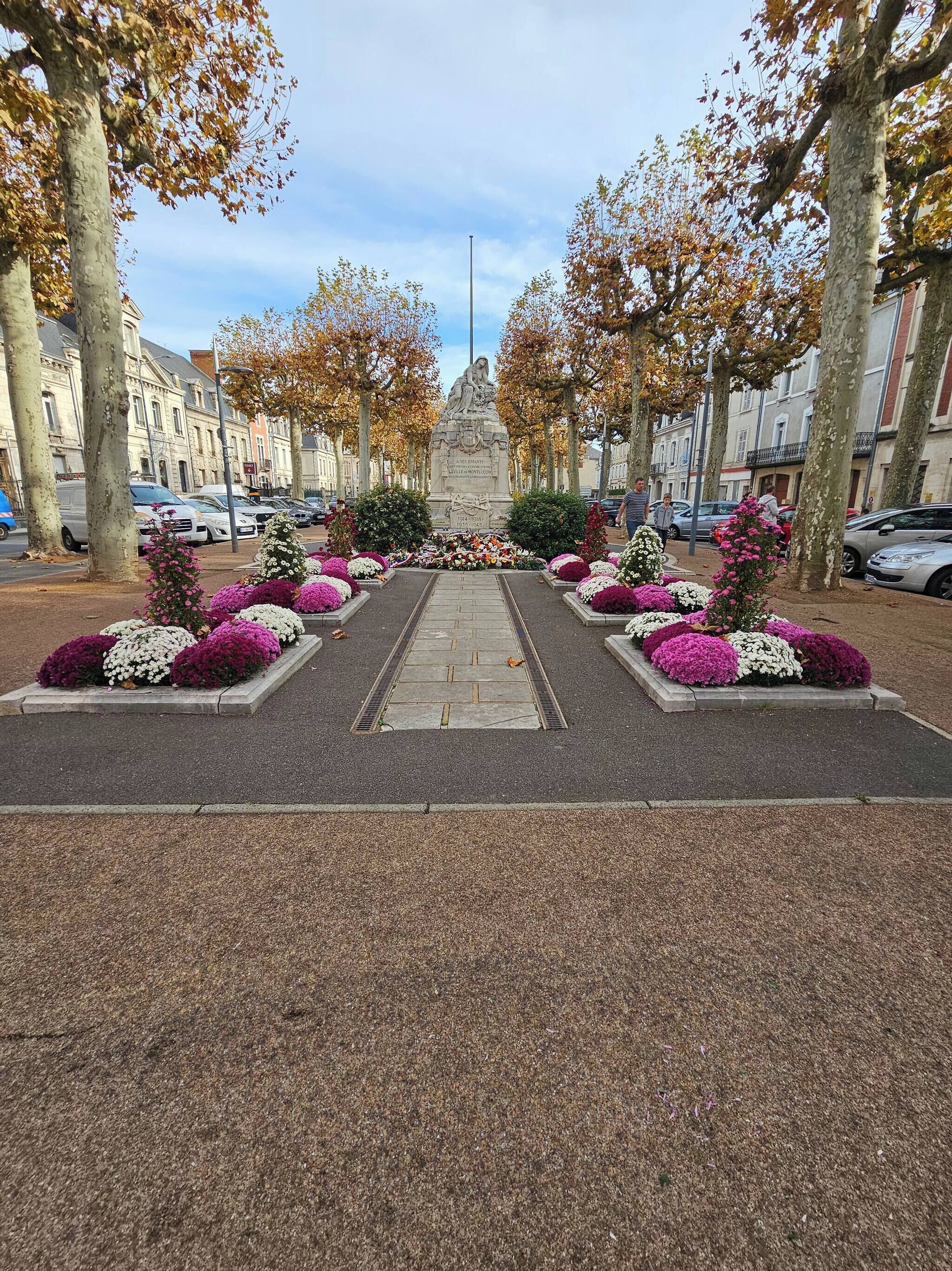 A memorial with a central statue, surrounded by symmetrical flower arrangements and a paved walkway.