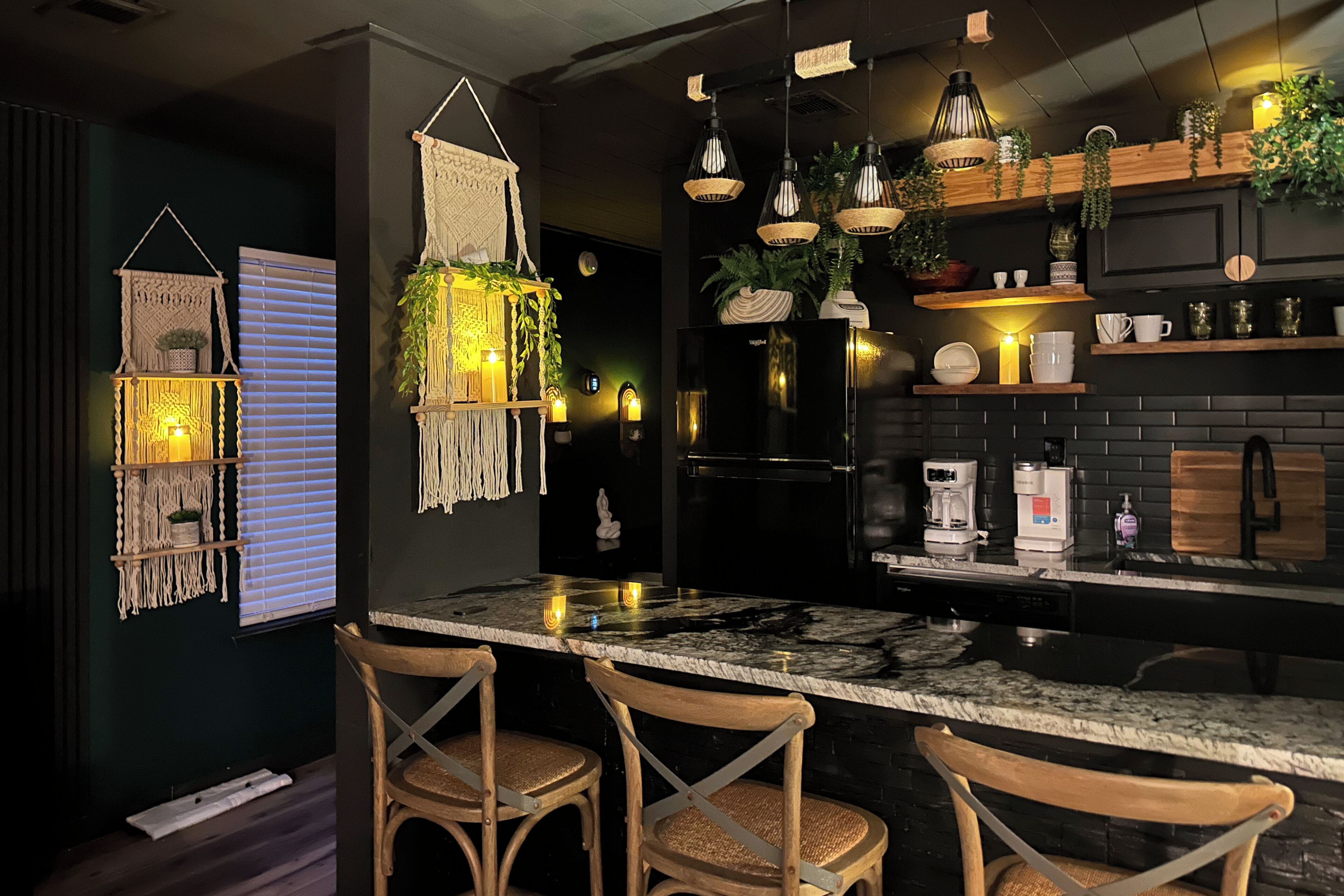 A modern kitchen with a marble countertop, wooden chairs, hanging plants, and a black refrigerator.