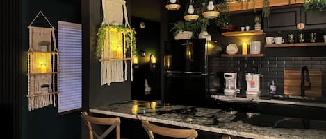 A modern kitchen with a marble countertop, wooden chairs, hanging plants, and a black refrigerator.