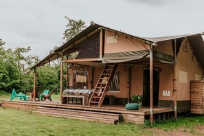 A two-story wooden cabin with a covered porch, outdoor seating, and a staircase leading to the upper level. - Kampari - Luxury Safari Tent - De Jungle (Delfstrahuizen)