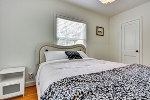 Een slaapkamer met een bed, nachtkastje, raam met zonwering, deur en een ingelijst schilderij aan de muur.
