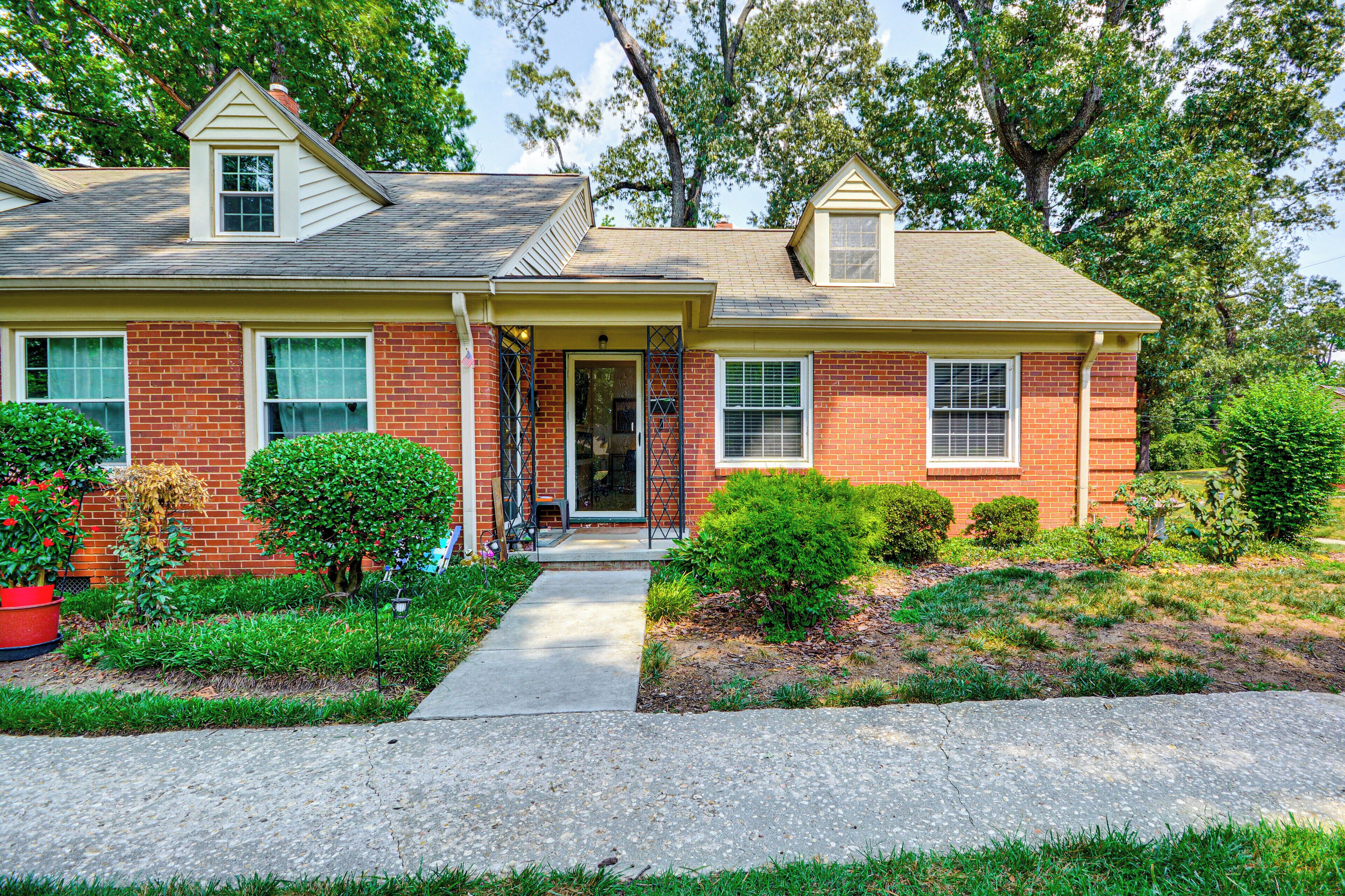 A two-story residential house with a front porch, brick exterior, and a small front yard with various plants.